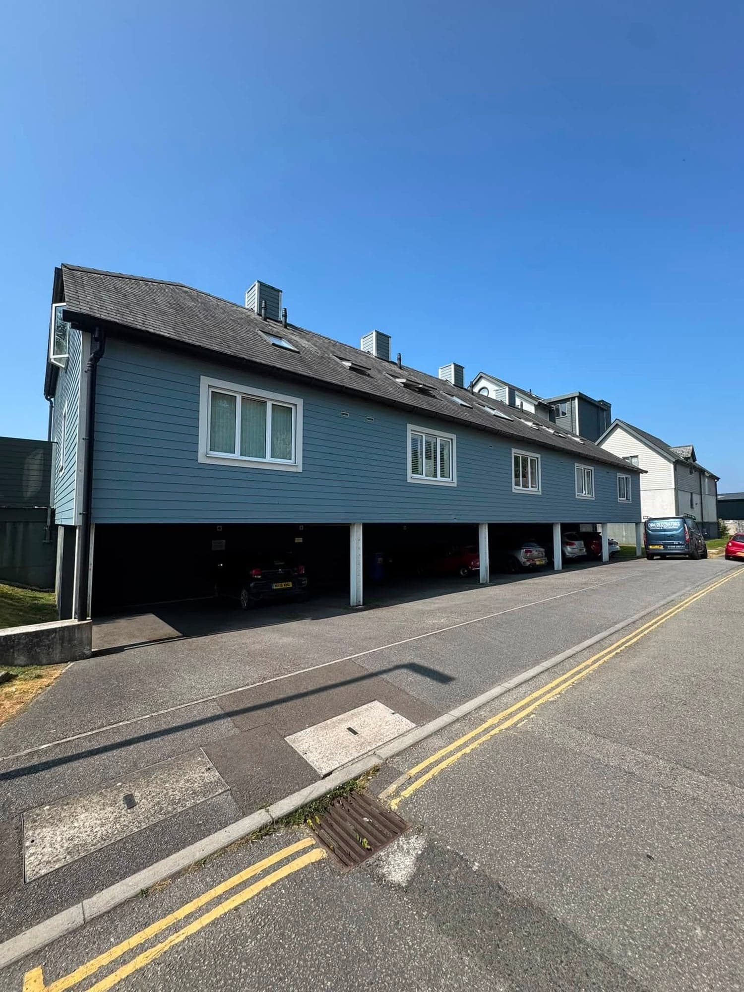 Long street elevation of a block of flats with pale blue painted timber cladding, dormer windows and parking under the building