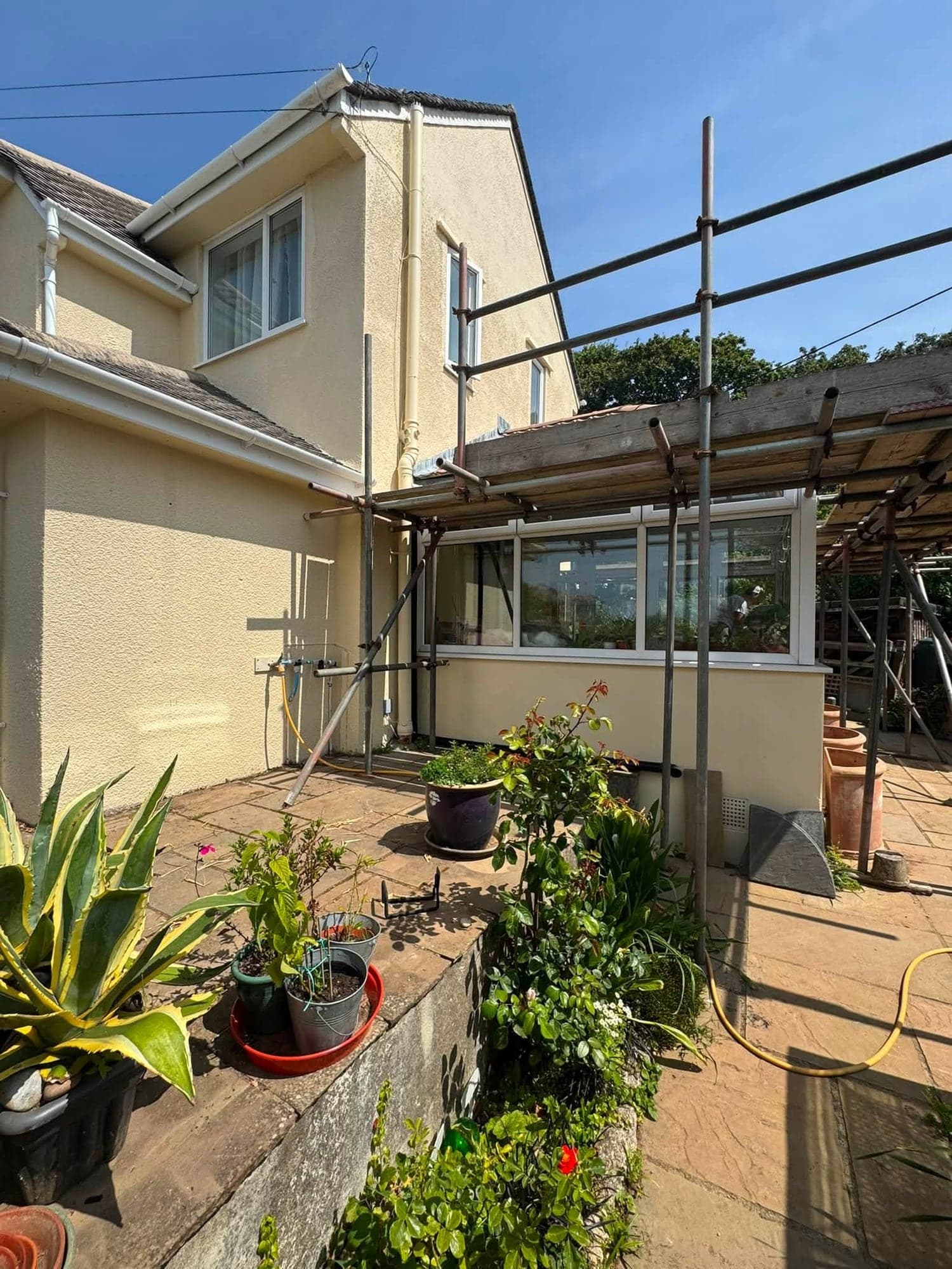 Rear of a cream rendered house with scaffolding over a conservatory during an exterior repaint, plants on a patio in the foreground