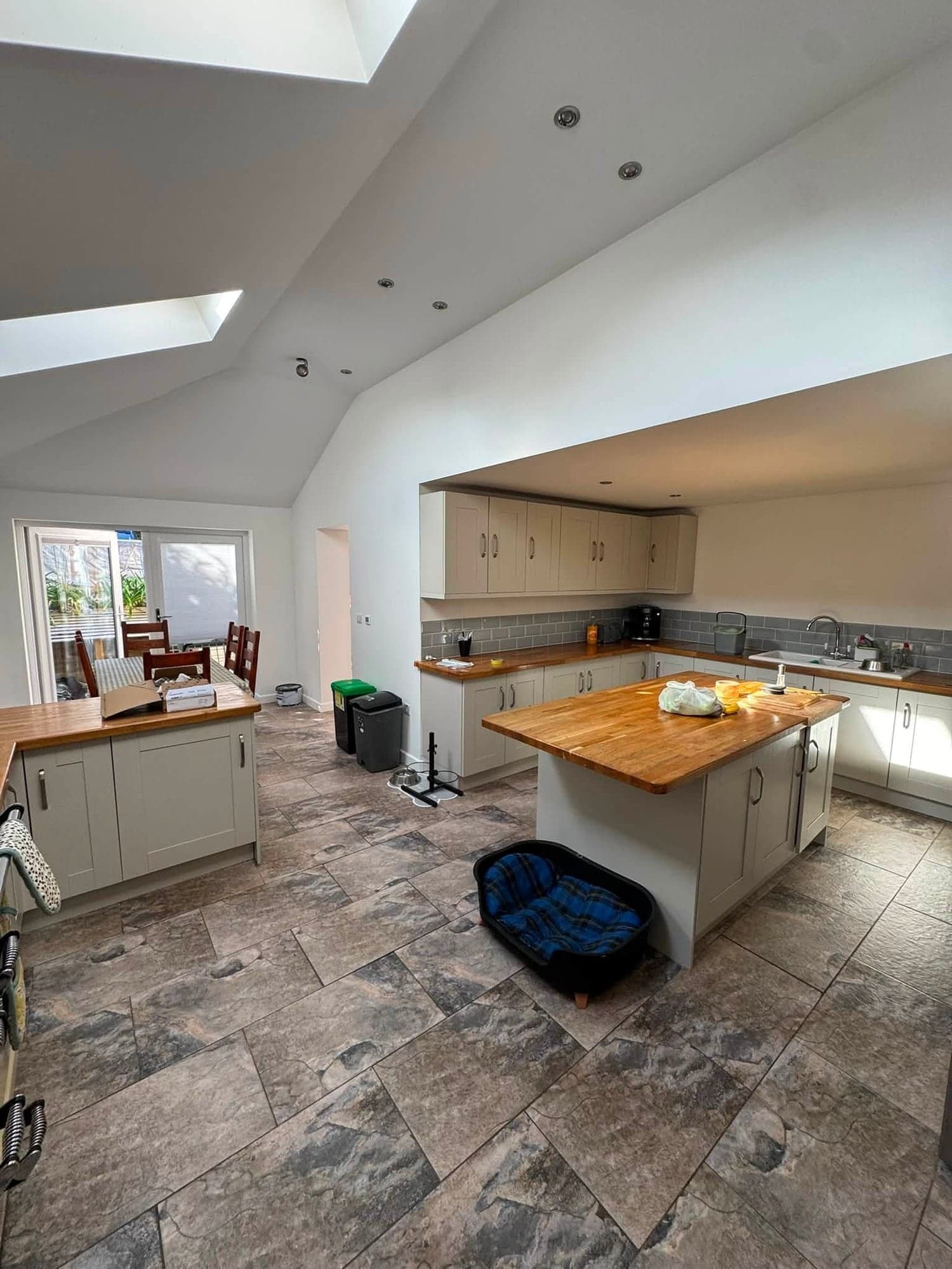 Open-plan kitchen diner with a white vaulted ceiling and rooflights, pale grey shaker units, oak worktops and a slate-effect floor