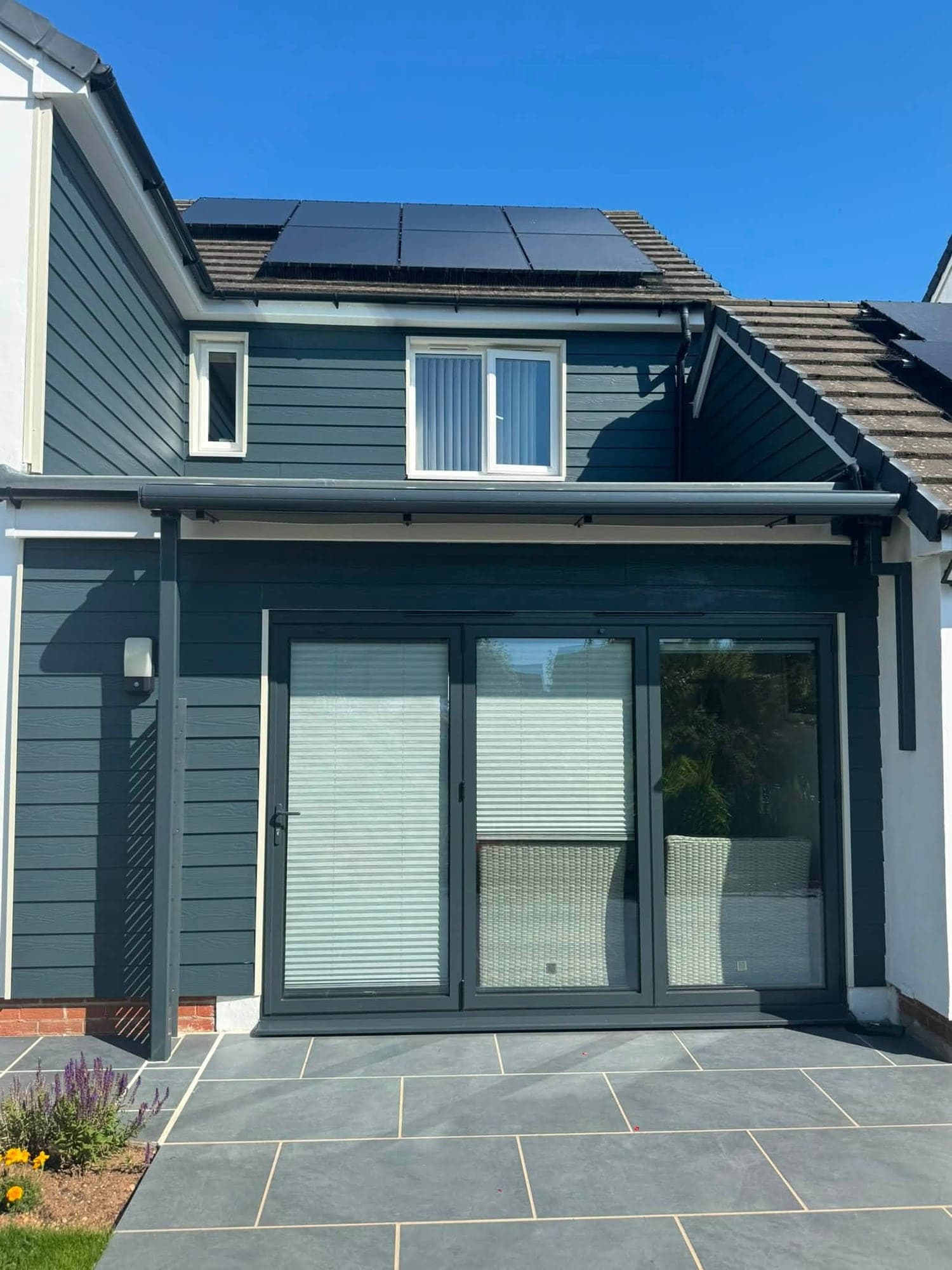 Rear of a house with dark navy painted cladding, grey bifold doors and a white rendered wall beside a grey slab patio