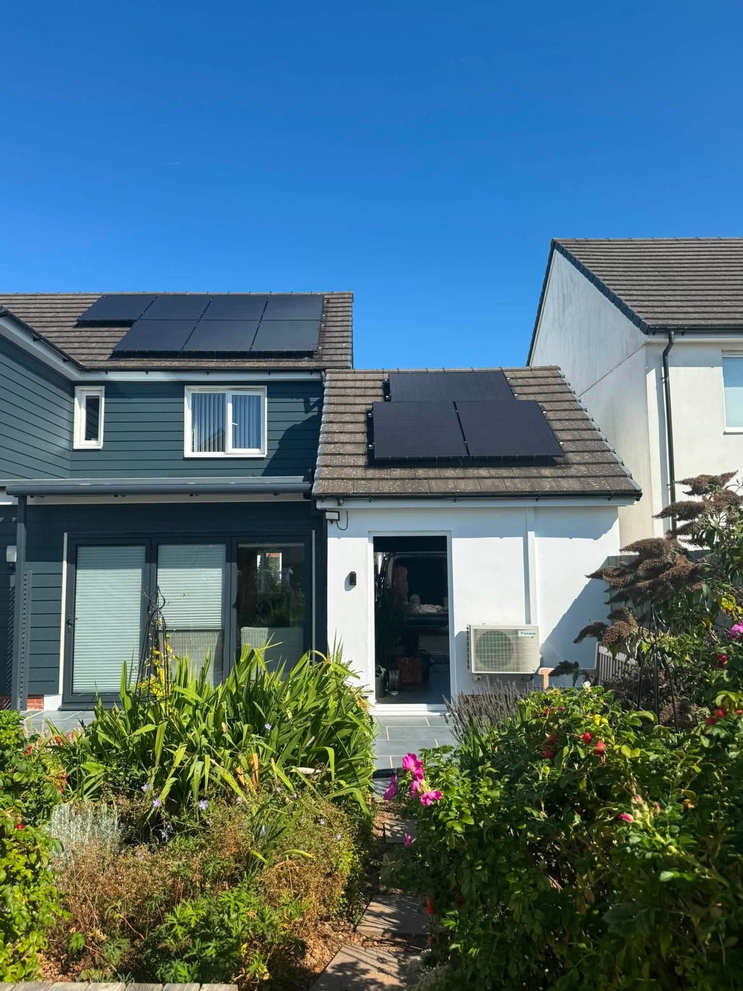 Rear of a house with navy cladding, a white rendered single-storey extension and solar panels on both roofs, with shrubs and roses in front