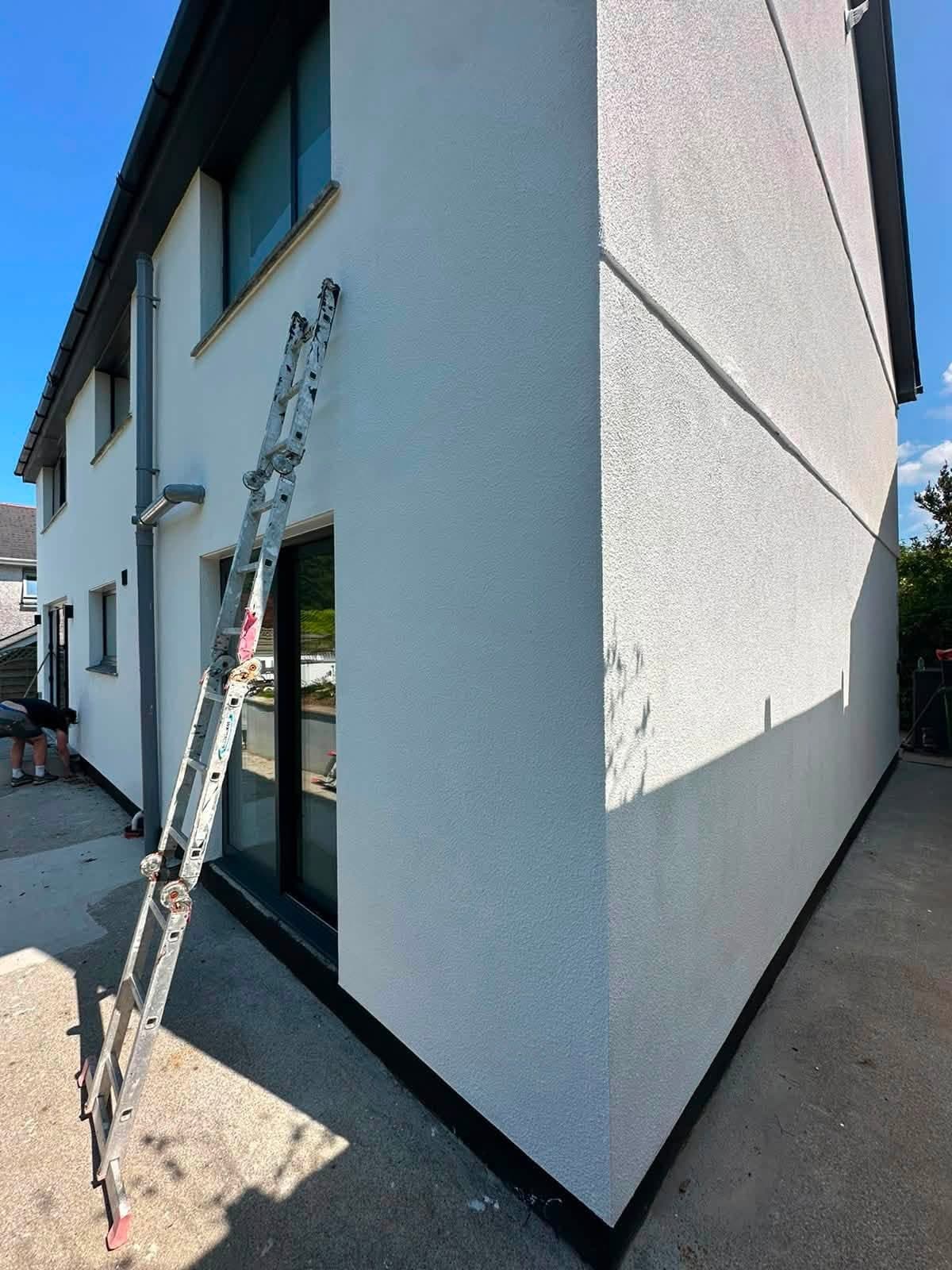 Ladder against a newly painted white rendered gable with dark grey window frames and a black plinth at ground level