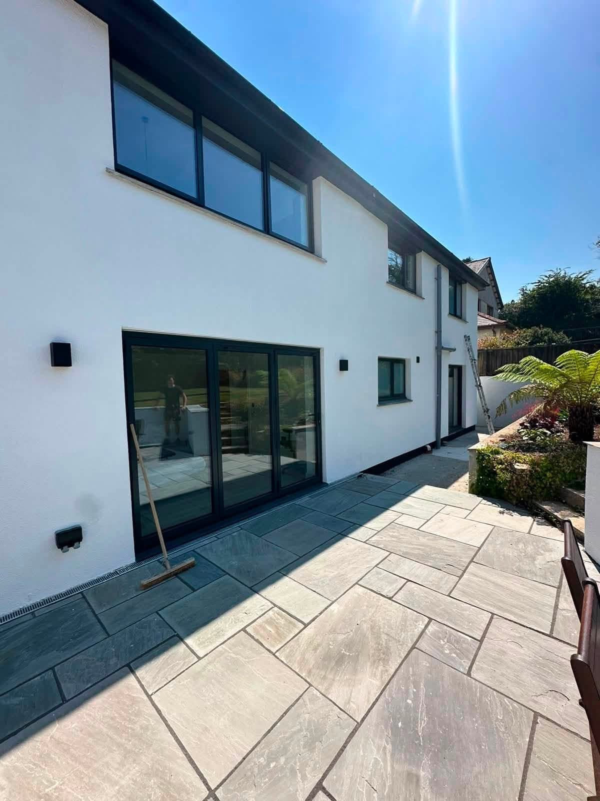 Rear elevation of a modern house in bright white render with dark bifold doors and windows, a slate patio and a ladder at the far end