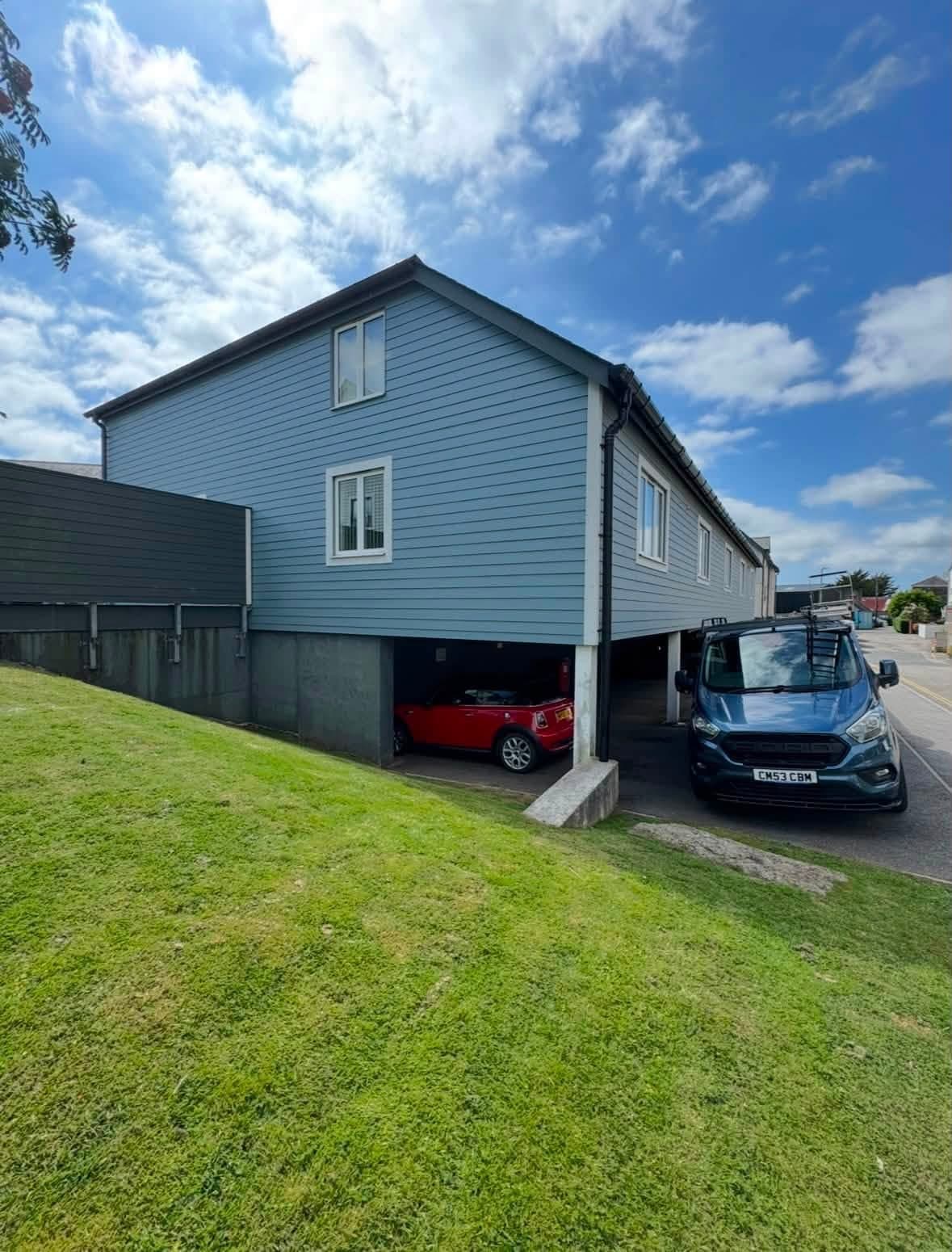 Gable end of a block of flats in pale blue timber cladding with white windows, cars parked in the undercroft beneath and a decorator's van beside it